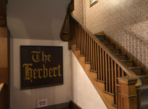A wooden staircase with a decorative wall and a sign reading "The Herbert" on the wall beside it.
