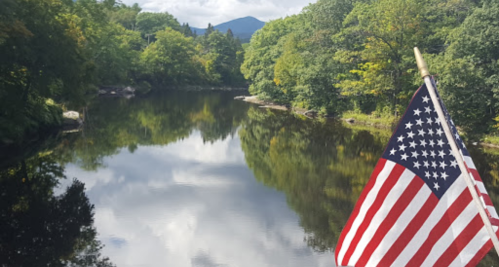 A serene river scene with lush green trees and a mountain in the background, featuring an American flag in the foreground.