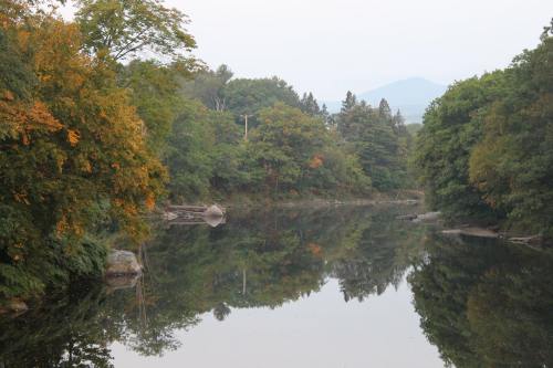A serene river surrounded by trees with autumn foliage, reflecting the landscape and mountains in the calm water.