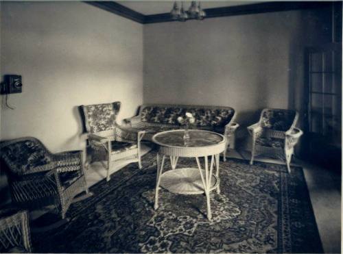 A vintage living room with wicker furniture, a round table, and a patterned rug, softly lit by natural light.