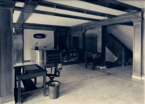A vintage interior of a lobby with wooden paneling, a reception desk, a staircase, and simple furniture.