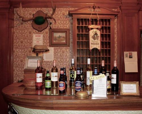 A wooden bar with various bottles of wine and beer, set against a patterned wall and a welcome sign.