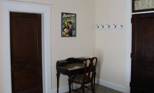 A small room featuring a desk, a chair, a poster on the wall, and a coat rack with hooks. Two doors are visible.