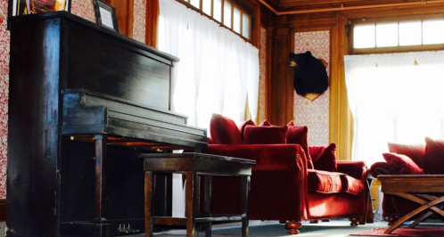 A cozy room featuring a black piano, red sofas, and a wooden coffee table, with large windows and decorative walls.