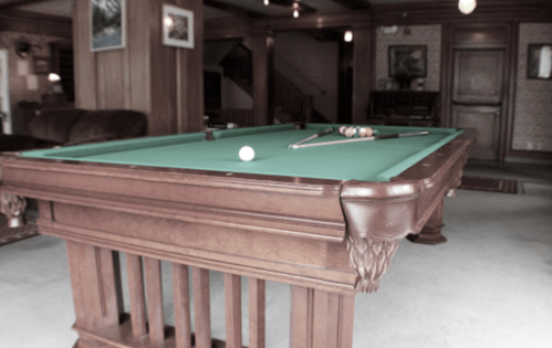 A vintage wooden pool table with a green felt surface, cue sticks, and a single white ball in a cozy room.