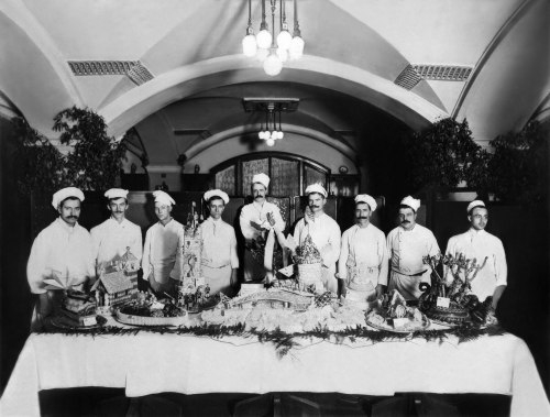 A historic black-and-white photo of chefs posing behind a lavishly decorated table of food in a grand dining hall.