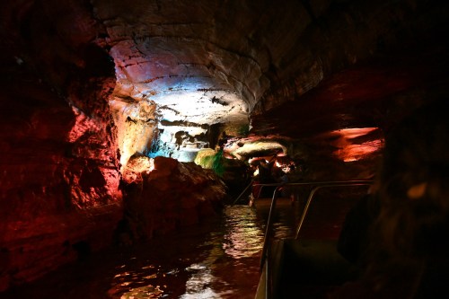 A colorful cave interior with illuminated rock formations and a calm waterway, viewed from a boat.