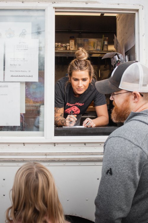 A woman at a food truck window takes an order from a man while a child watches nearby.