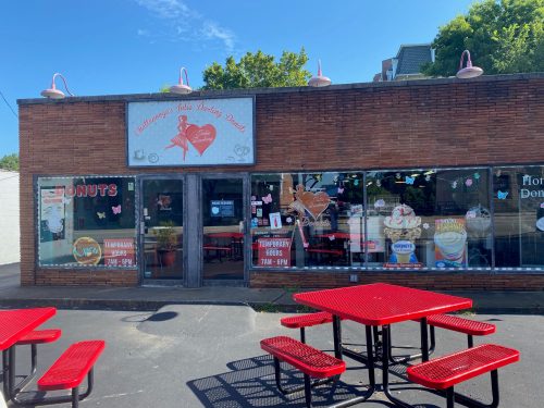 A brick donut shop with a red outdoor seating area, featuring large windows and colorful signage.