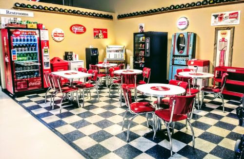 A retro diner setup with red chairs and tables, vintage Coca-Cola decor, and a soda vending machine.