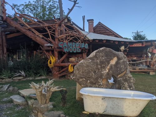 A rustic cabin with a sign reading "Campfire Woods," featuring a stone sculpture and an old bathtub in the foreground.