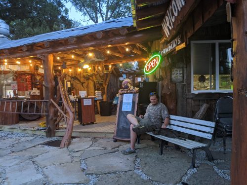 A man sits on a bench outside a rustic, illuminated building with an "Open" sign, surrounded by trees.