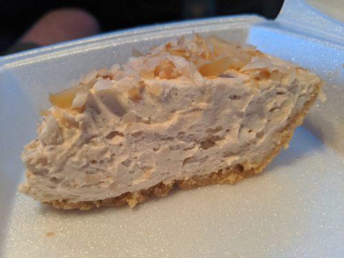 A slice of creamy cheesecake topped with shredded coconut, resting in a white takeout container.