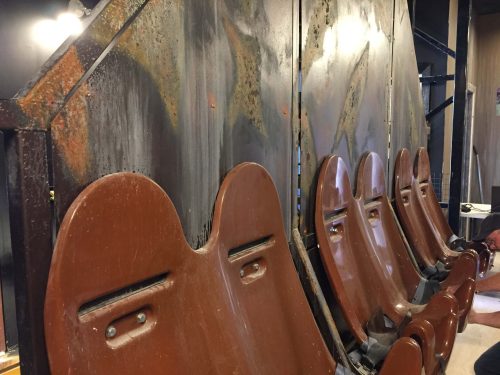 A row of worn, brown seats against a weathered metal wall with rust and peeling paint.