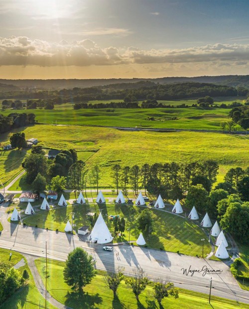 Aerial view of teepees arranged in a green landscape, with sunlight streaming through clouds in the background.