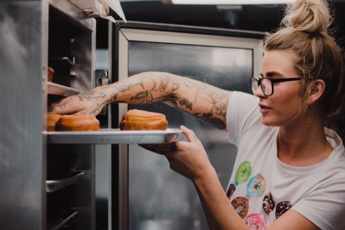 A woman with tattoos and glasses places donuts on a tray in a bakery.