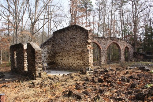 Ruins of a stone structure surrounded by trees, with remnants of arches and a sign in a forested area.