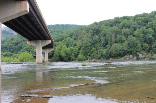 A bridge spans over a calm river, surrounded by lush green trees and hills under a cloudy sky.