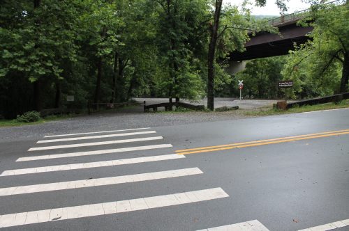 A quiet road with a crosswalk, surrounded by trees, leading to a gravel parking area under a bridge.