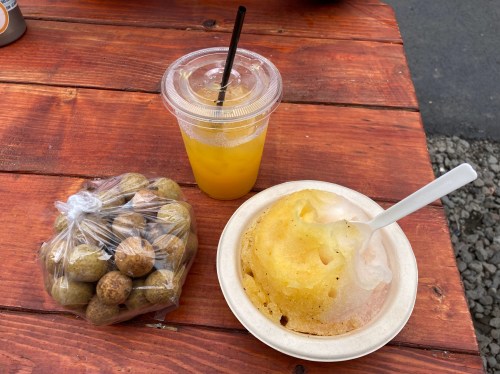 A cup of orange drink, a bag of small round fruits, and a bowl of shaved ice with syrup on a wooden table.