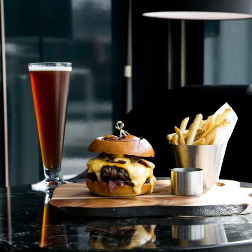 A gourmet cheeseburger with fries and a tall glass of dark beer on a wooden platter, set on a stylish table.