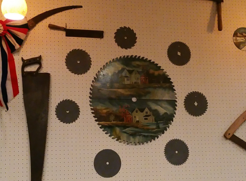 A collection of vintage saws and circular blades displayed on a pegboard, featuring a painted scene of houses.