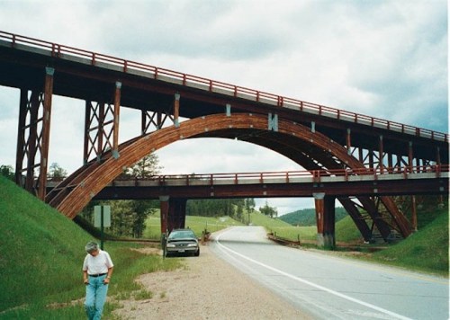 A unique wooden bridge arches over a road, with a person walking nearby and a car parked on the side.