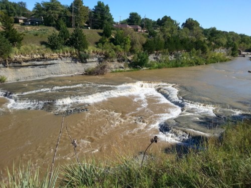 A river flows over rocky terrain, creating small waterfalls, surrounded by greenery and residential homes in the background.