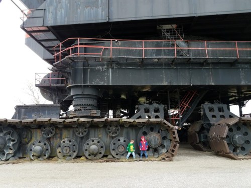 Two children stand beside a large, industrial machine with massive tracks, showcasing its size and structure.