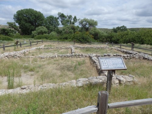 Ruins of a stone foundation surrounded by grass, with a sign detailing the site's history in a natural landscape.