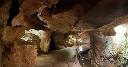 A dimly lit cave interior with rocky walls and a pathway bordered by a chain railing.