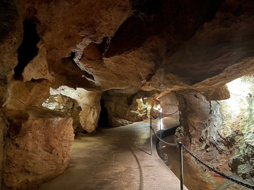 A dimly lit cave interior with rocky walls and a winding path bordered by a chain railing.