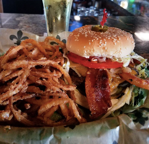 A juicy burger with bacon, cheese, lettuce, and tomato, served with crispy onion strings and a drink in the background.