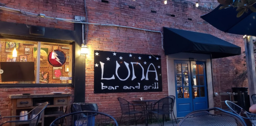 Exterior of Luna Bar and Grill, featuring a brick wall, a large sign, and outdoor seating under string lights.