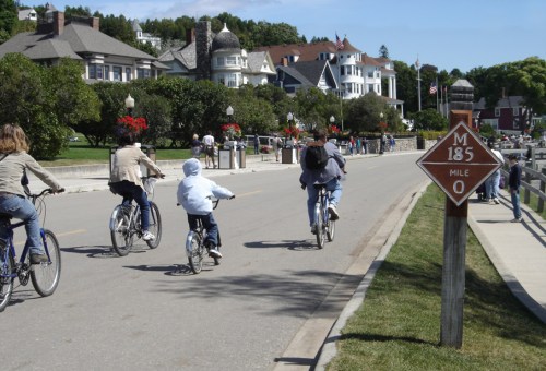 Bicyclists ride along a scenic road with historic homes and a mile marker sign in a vibrant, sunny setting.