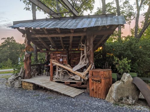 A rustic outdoor structure with a wooden roof, surrounded by trees, featuring seating and a trash bin nearby.