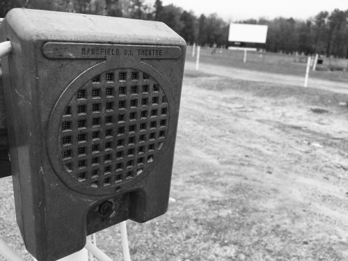 A vintage drive-in speaker stands in the foreground, with a blurred movie screen in the background on a cloudy day.