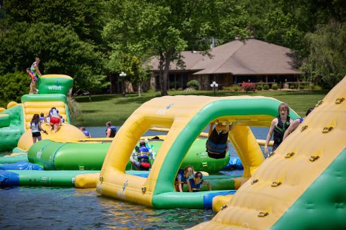 Colorful inflatable water park with people playing and climbing on structures in a lake setting. Green trees in the background.
