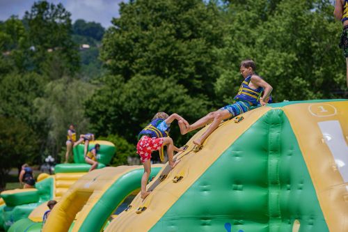 Two children climb an inflatable water slide, surrounded by greenery and other kids enjoying the water park.