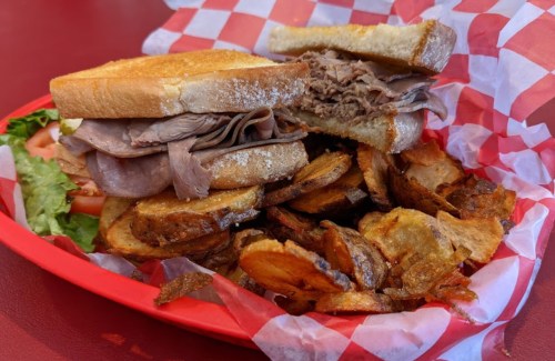 A hearty sandwich with roast beef on toasted bread, served with a side of crispy potato wedges in a red basket.