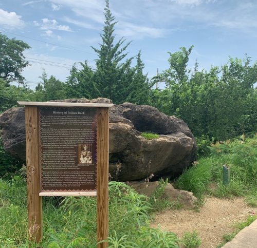 A wooden sign about the history of Indian Rock stands beside a large rock formation, surrounded by greenery and blue sky.