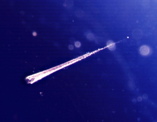 A bright, elongated comet streaks across a deep blue sky, leaving a faint trail behind it.