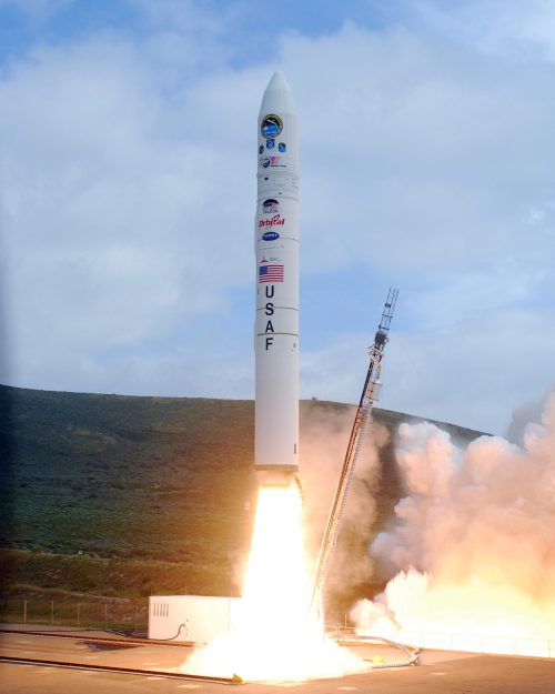 A rocket launches from a pad, with flames and smoke billowing from its base against a cloudy sky.