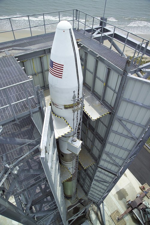 A rocket is positioned vertically in a launch pad structure, with the ocean visible in the background.