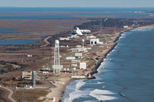 Aerial view of a coastal facility with buildings, a water tower, and a launch pad along the shoreline and marshland.