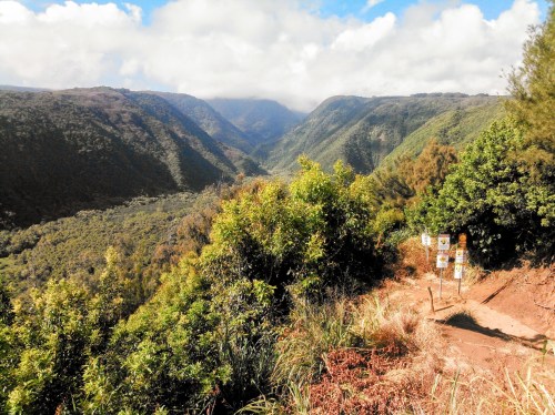 A scenic view of a lush valley surrounded by mountains under a partly cloudy sky, with trail signs in the foreground.