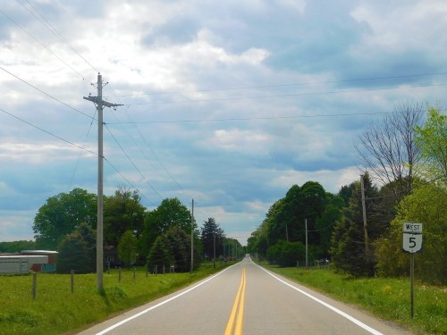 A rural road stretches into the distance, flanked by trees and power lines under a cloudy sky. A highway sign indicates "West 5."