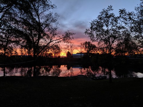 A serene sunset over a calm pond, framed by trees, with vibrant orange and purple hues in the sky.