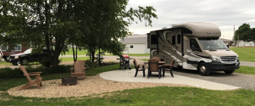 An RV parked beside a patio with chairs and a grill, surrounded by grass and trees.