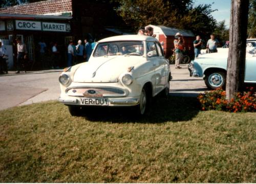 A vintage white car parked on grass in front of a market, with people gathered nearby.